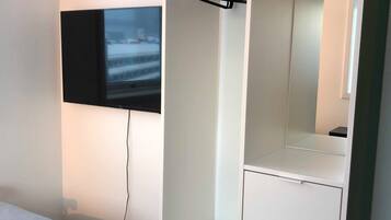 A bedroom with a bed, a TV mounted on the wall, a wardrobe with a mirror, and a shelf unit.