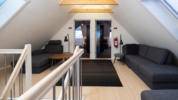 Un dormitorio estilo loft moderno con techo inclinado, vigas de madera integradas, un sofá gris, una mesita pequeña y una alfombra negra.