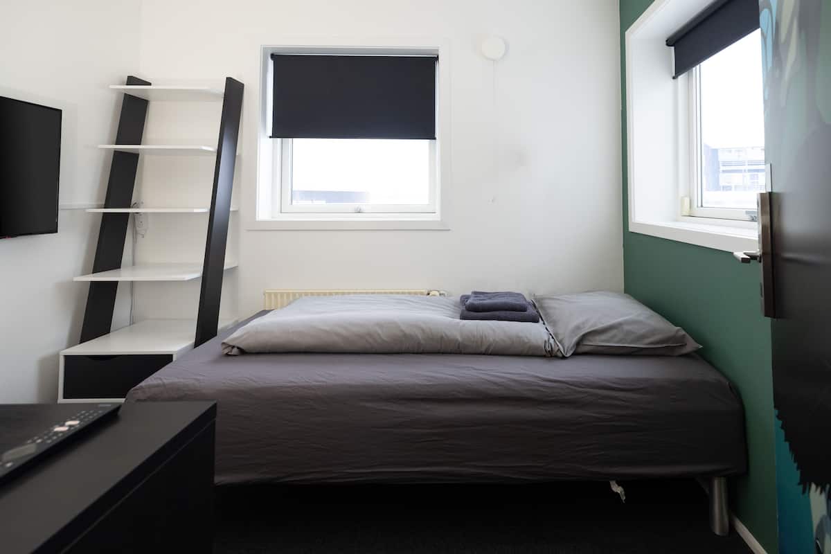 A small, neatly arranged bedroom with a bed, a shelf unit, a desk, and a window.