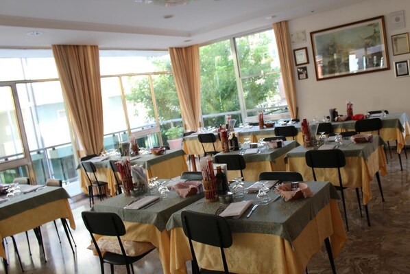A dining area with multiple tables set for service, large windows with curtains, and framed pictures on the walls.