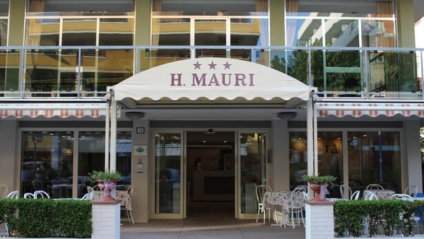 H. Mauri-hotellets fasad med en framträdande entré, glasdörrar och ett utomhusområde med sittplatser.
