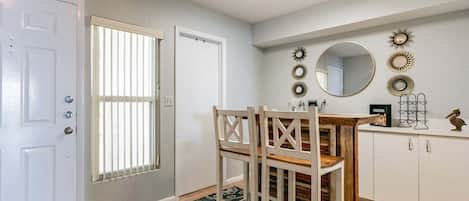 Een eetruimte met een tafel en stoelen, een witte deur, een raam met zonwering en een plafondlamp.