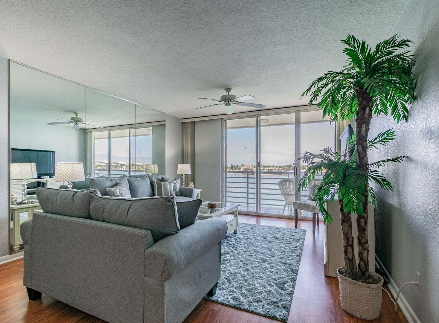 Een moderne woonkamer met een grijze bank, een grote spiegel en uitzicht op zee door een glazen schuifdeur.