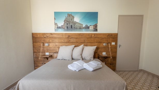 A hotel room with a wooden headboard, a bed with a beige bedspread, two pillows, and a folded towel on the bed. There is a painting of a building on the wall above the bed. - Violet Guest House (Syracuse)