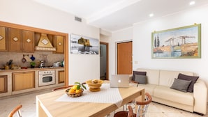 A modern kitchen with wooden cabinets, a dining table with chairs, a laptop, and a sofa in a living area with a painting on the wall.