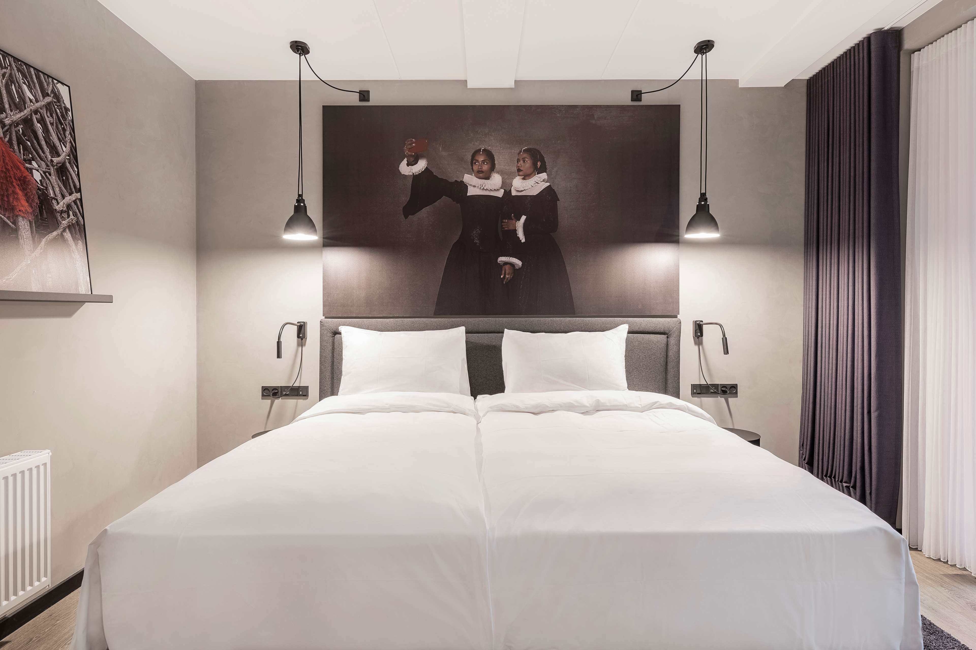 A modern hotel room with a large bed, two bedside tables, a headboard, a painting on the wall, and a window with curtains.