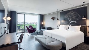 Et moderne hotelværelse med en stor seng, et skrivebord, en stol og udsigt over byen fra vinduet.
