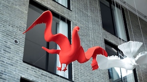 A red and white sculpture of a bird-like creature hanging from a building.