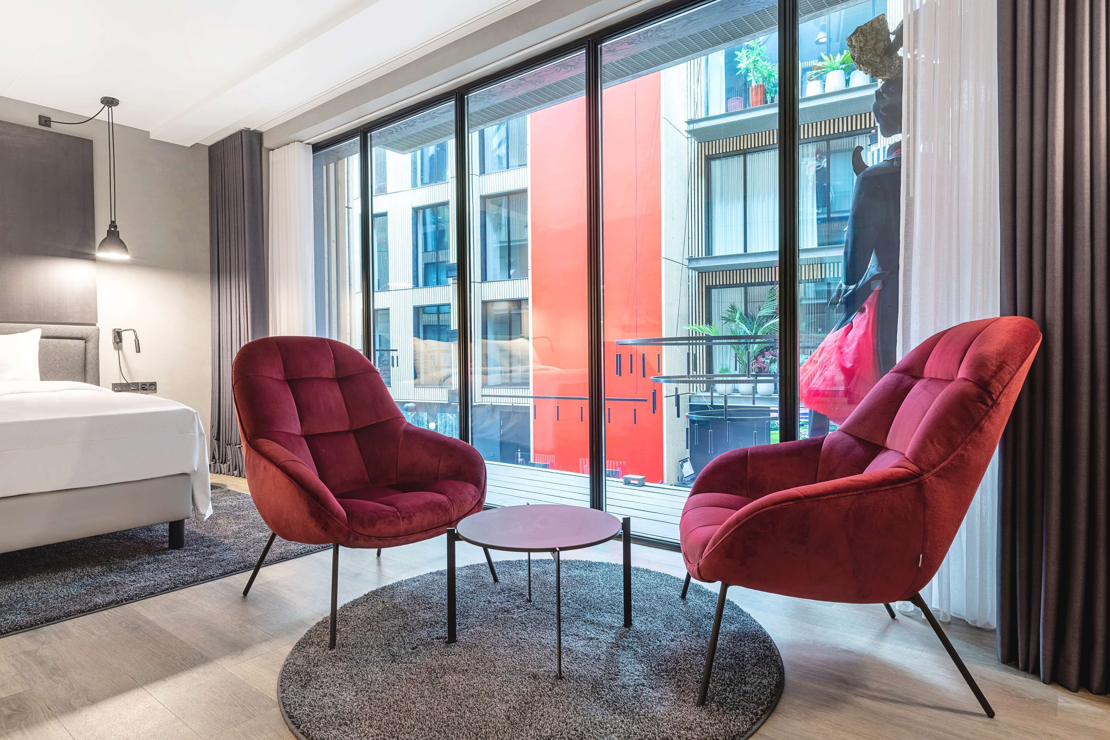 A modern hotel room with a large window, a bed, two red armchairs, and a small round table.