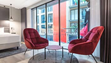 A modern hotel room with a large window, a bed, two red armchairs, and a small round table.