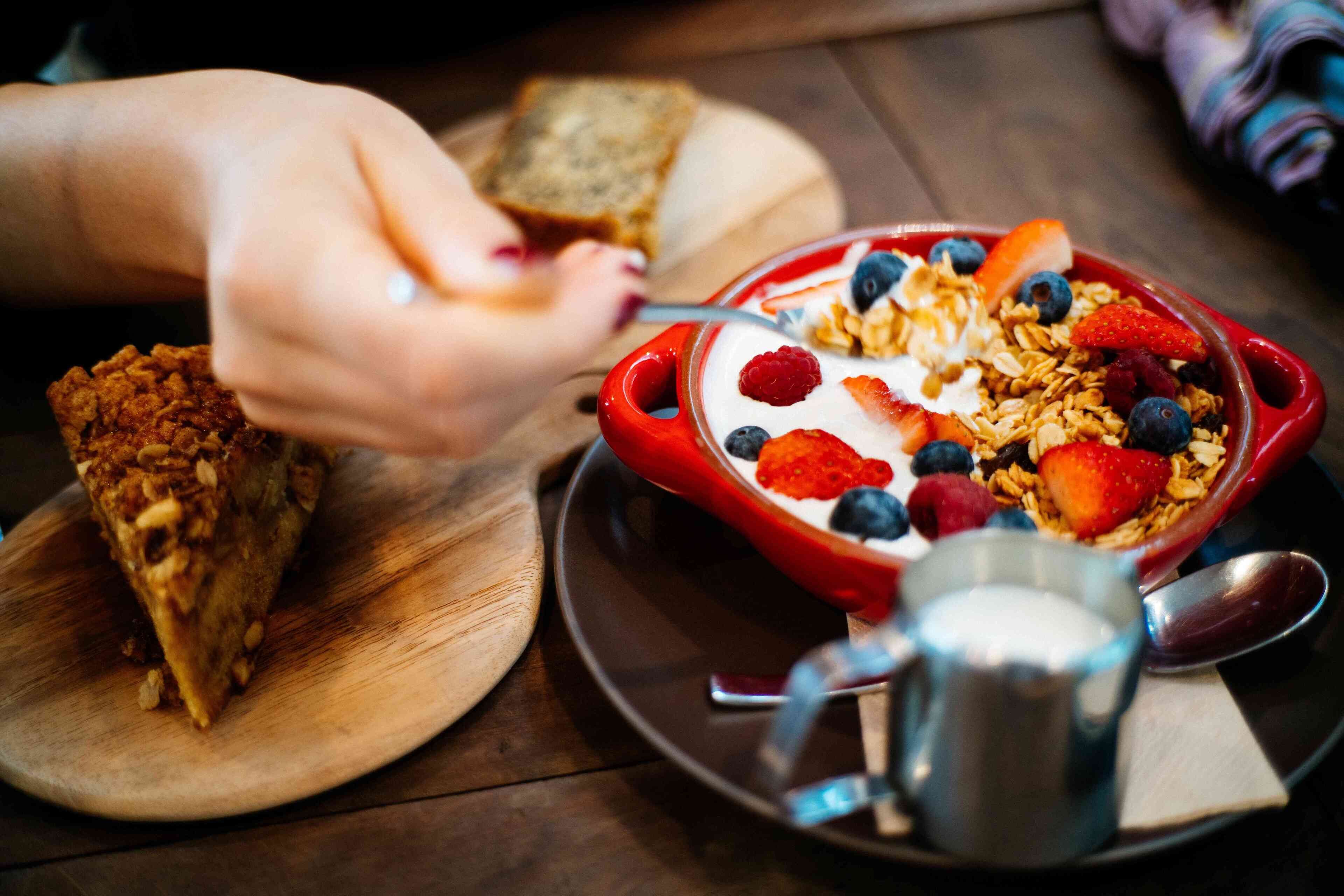 A breakfast setting with a bowl of cereal topped with fruits and yogurt, a slice of toast, and a metal cup on a wooden table.