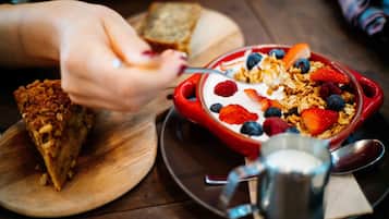 A breakfast setting with a bowl of cereal topped with fruits and yogurt, a slice of toast, and a metal cup on a wooden table.