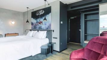 A modern hotel room with a large bed, a red chair, a painting, and a view of the city.