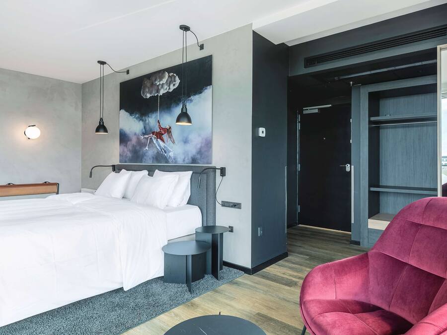 A modern hotel room with a large bed, a red chair, a painting, and a view of the city.