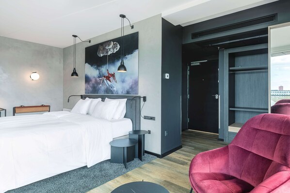 Habitación de hotel moderna con una cama grande, una silla roja, un cuadro y vistas a la ciudad.