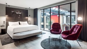 A modern hotel room with a large bed, two red armchairs, a small table, and a large window offering a view of the city. - Radisson RED Aarhus (Aarhus)
