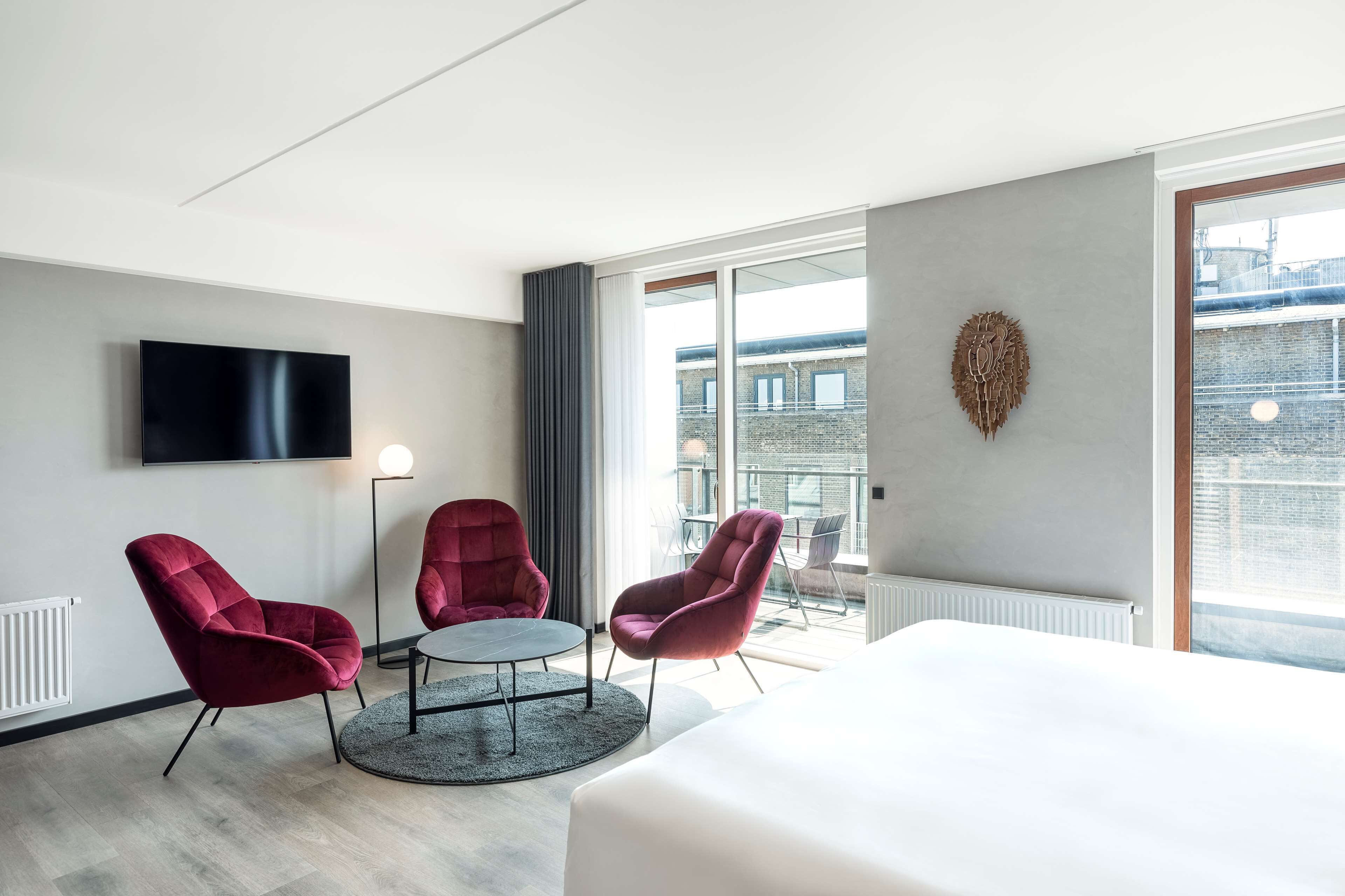 Una habitación de hotel moderna con una cama grande, dos sillones rojos, una mesa redonda, un televisor de pantalla plana y un ventanal con vistas a la ciudad.