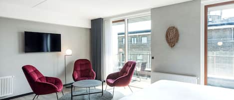 Una habitación de hotel moderna con una cama grande, dos sillones rojos, una mesa redonda, un televisor de pantalla plana y un ventanal con vistas a la ciudad.