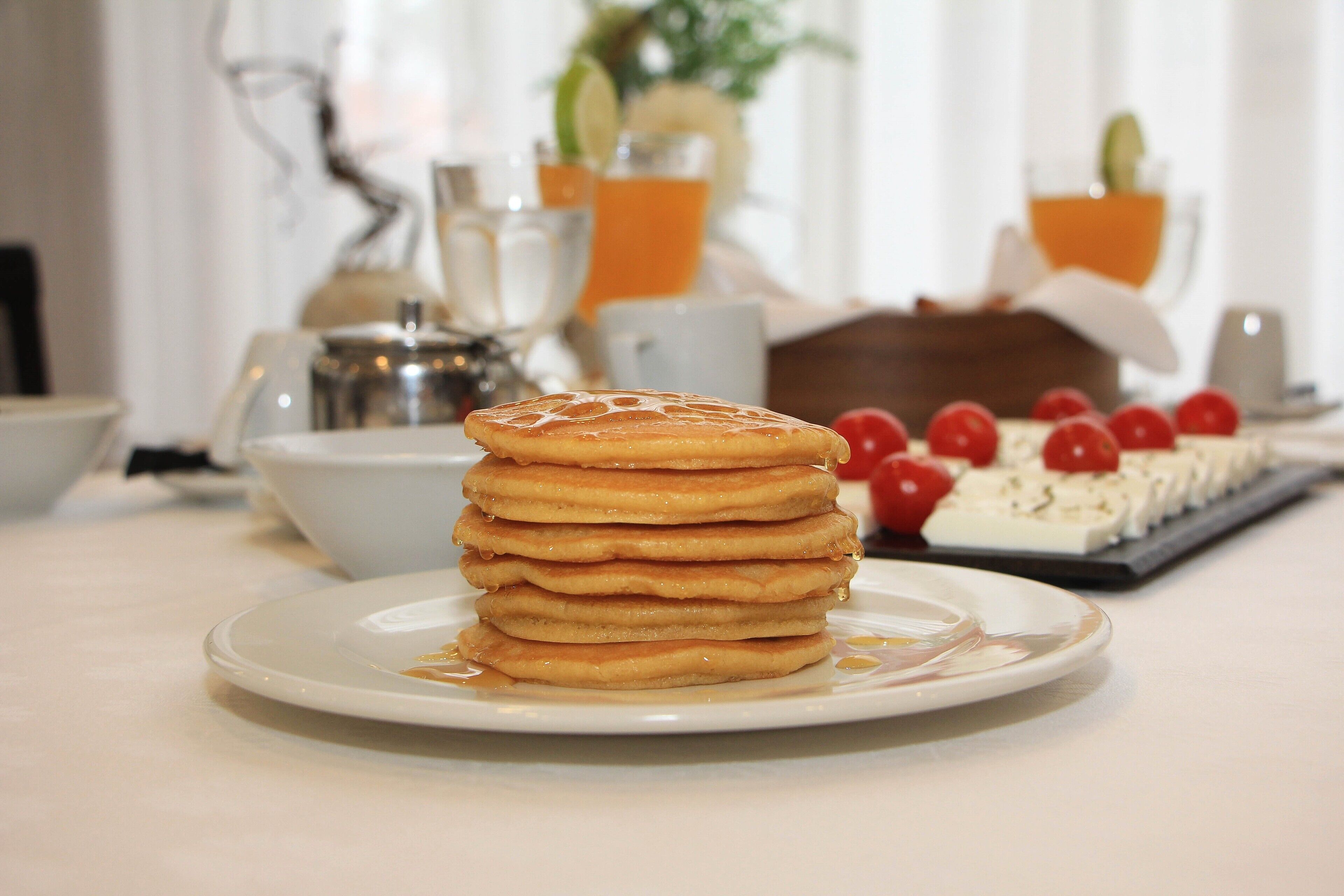 Une pile de crêpes sur une assiette, nappée de sirop, un dessert aux cerises et une cafetière en arrière-plan.