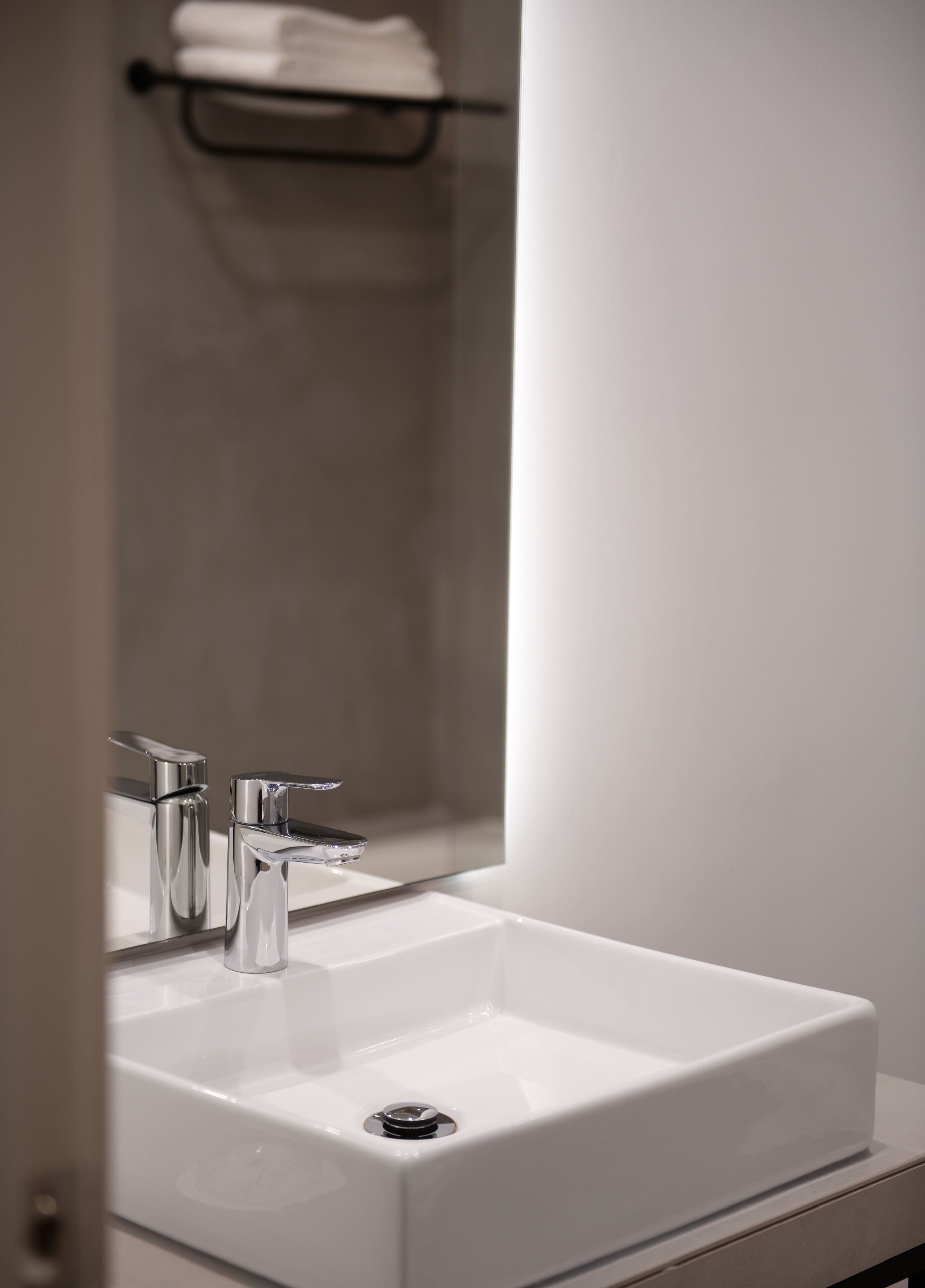 Un baño con un lavamanos blanco, grifo cromado y un espejo que refleja un perchero.