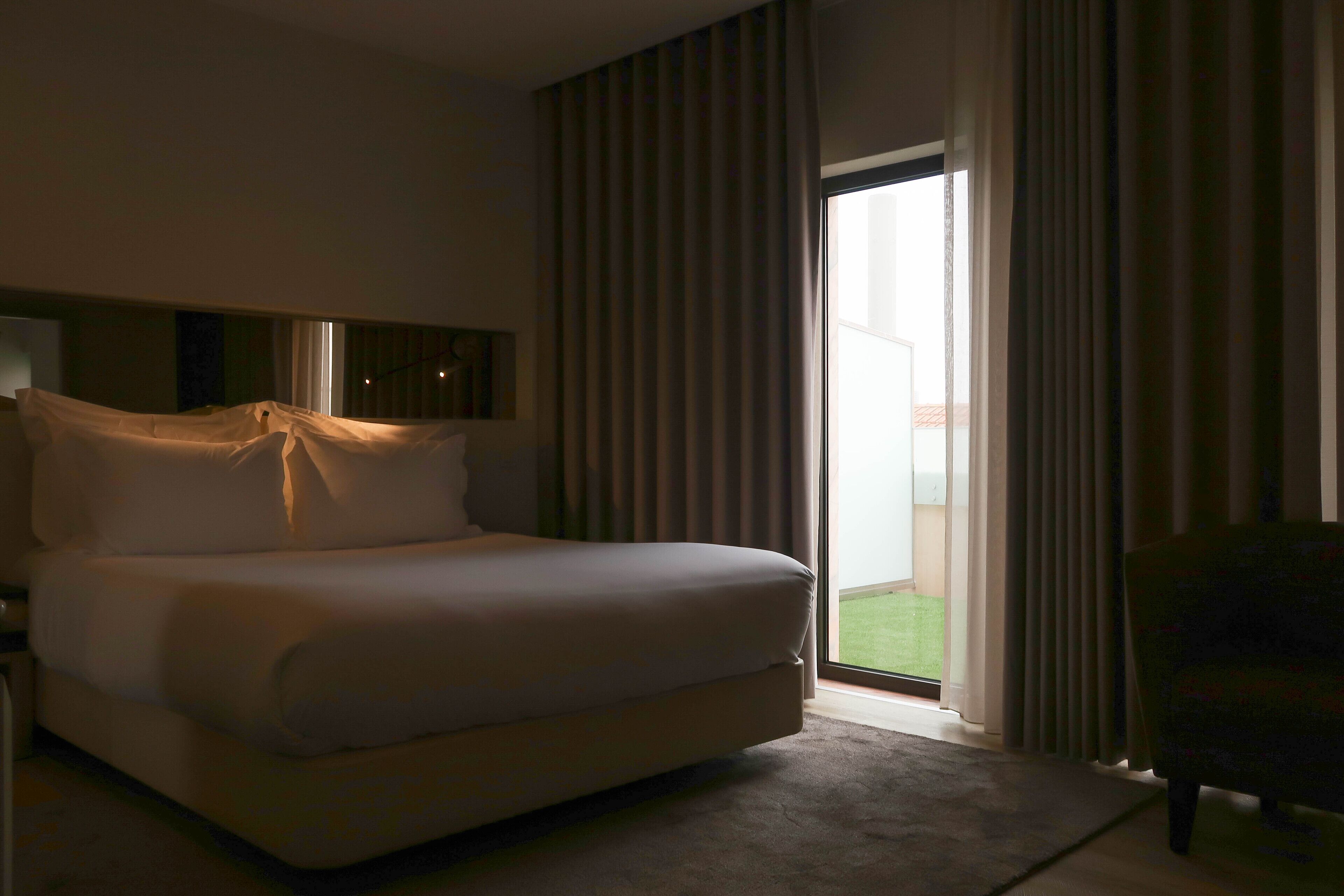 Ein Hotelzimmer mit einem großen Bett, einem Schreibtisch, einem Stuhl und Blick ins Freie durch eine Schiebetür.