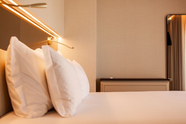 A neatly made bed with white linens and pillows, a bedside table, a lamp, and a mirror on the wall.