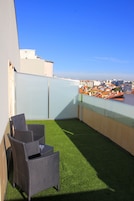 Una terraza en la azotea con césped artificial, dos sillones de mimbre y vistas a un paisaje urbano.