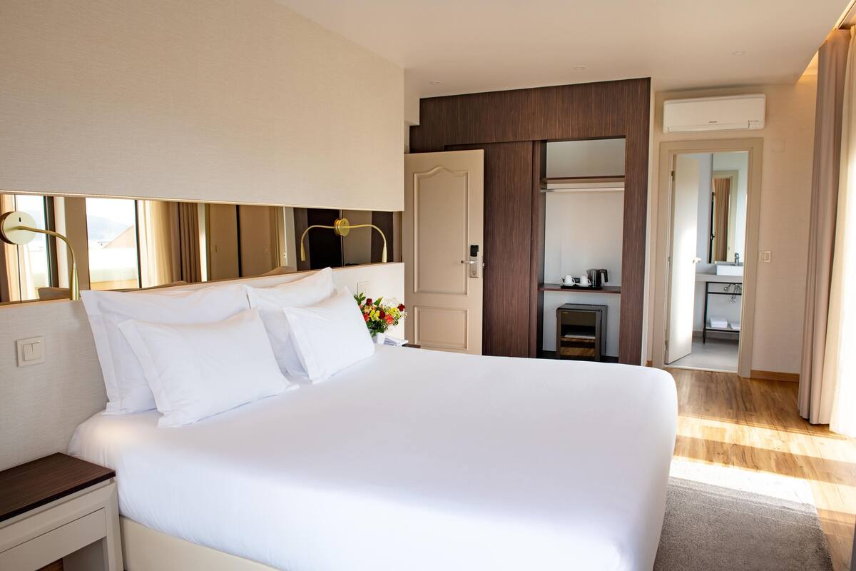 A hotel room with a large bed, bedside table, and a small desk with a chair. There is a mirror above the bed and a door leading to a bathroom.