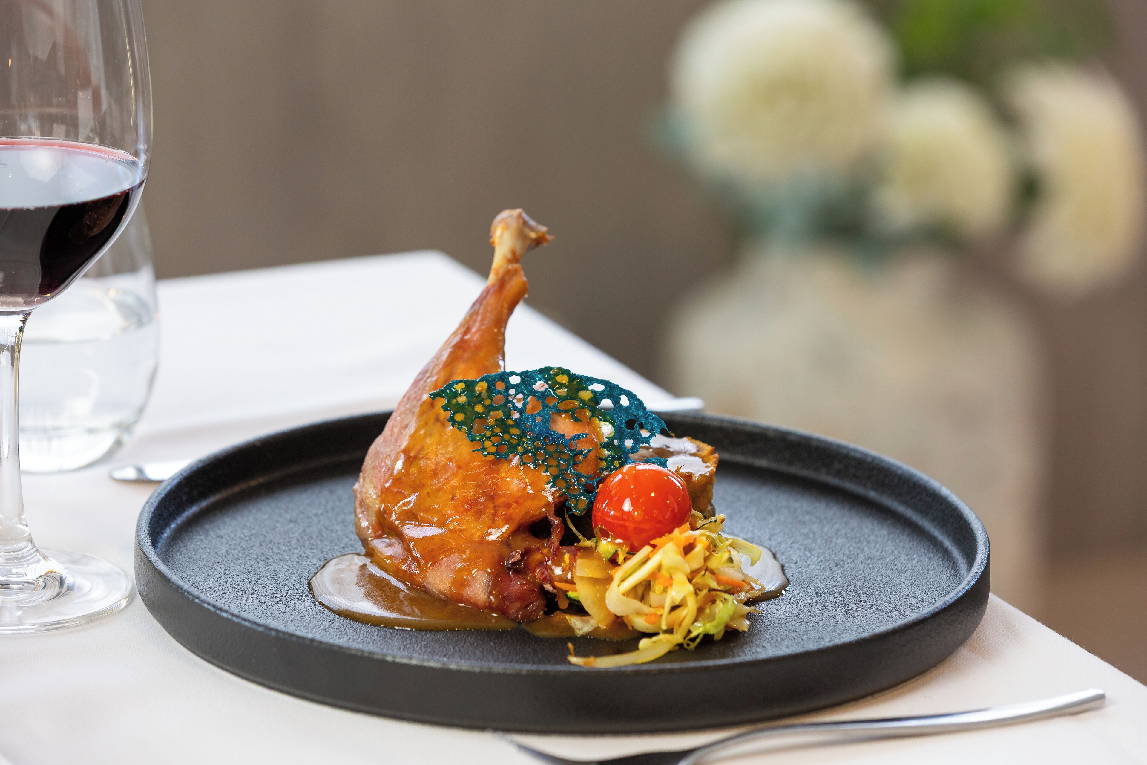 A plate with a roasted duck, vegetables, and a cherry on a black plate.
