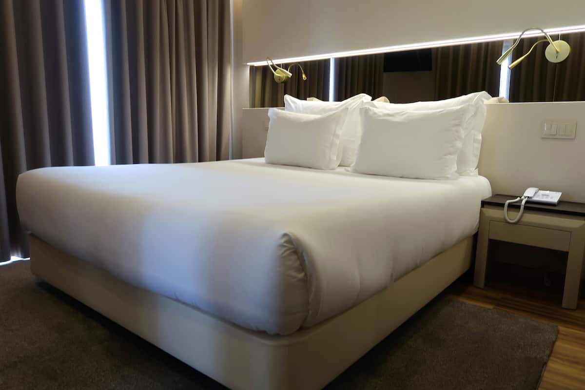 A neatly made bed with white linens, a bedside table with a phone, and a wall-mounted light fixture.