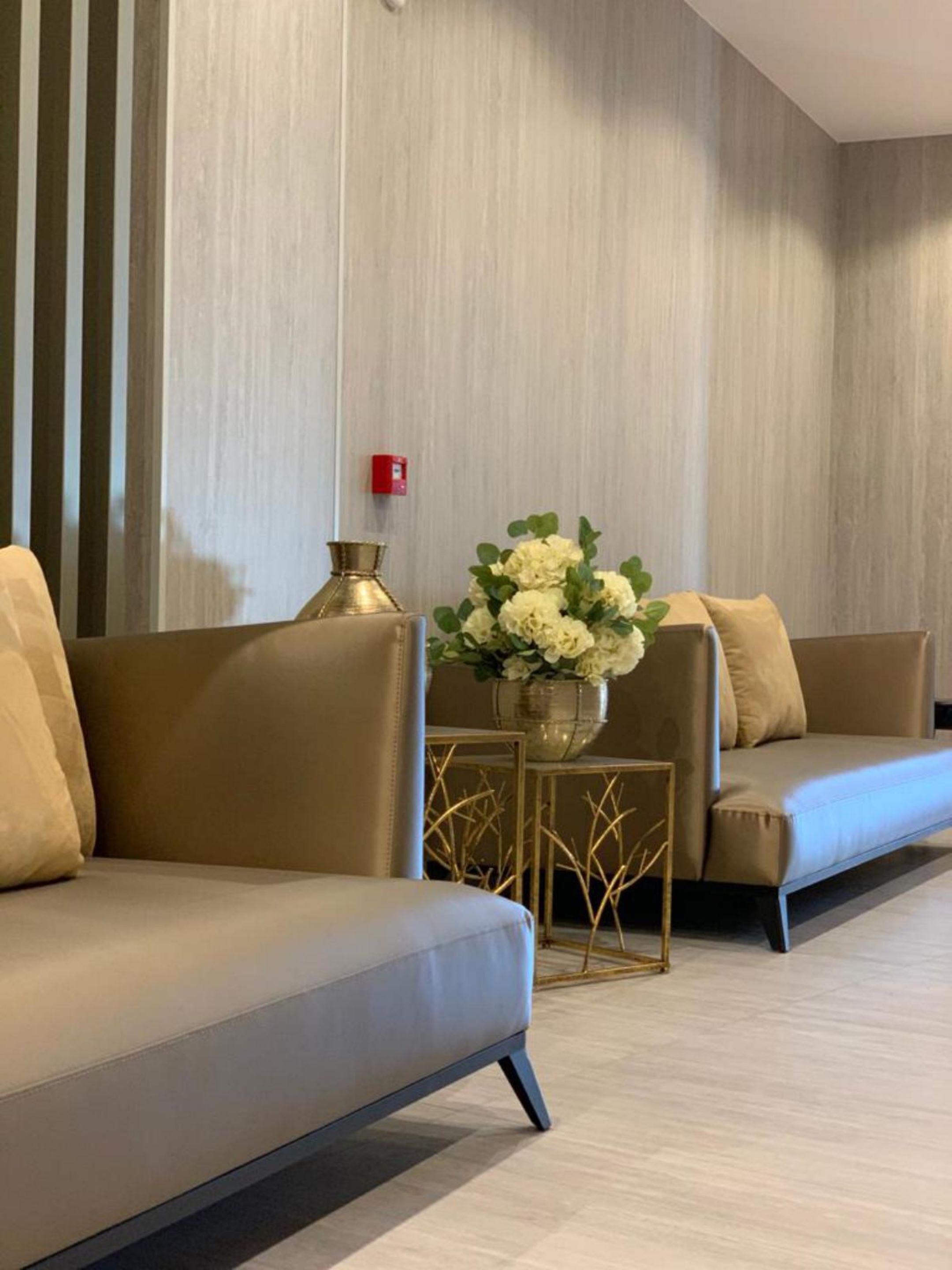 A modern hotel lobby with two sofas, a decorative table with a vase of flowers, and a fire alarm on the wall.