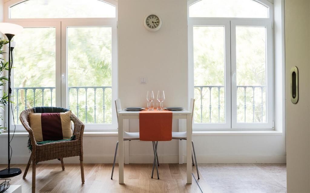 A room with a dining table set for two, a wicker chair, and a view through large windows.