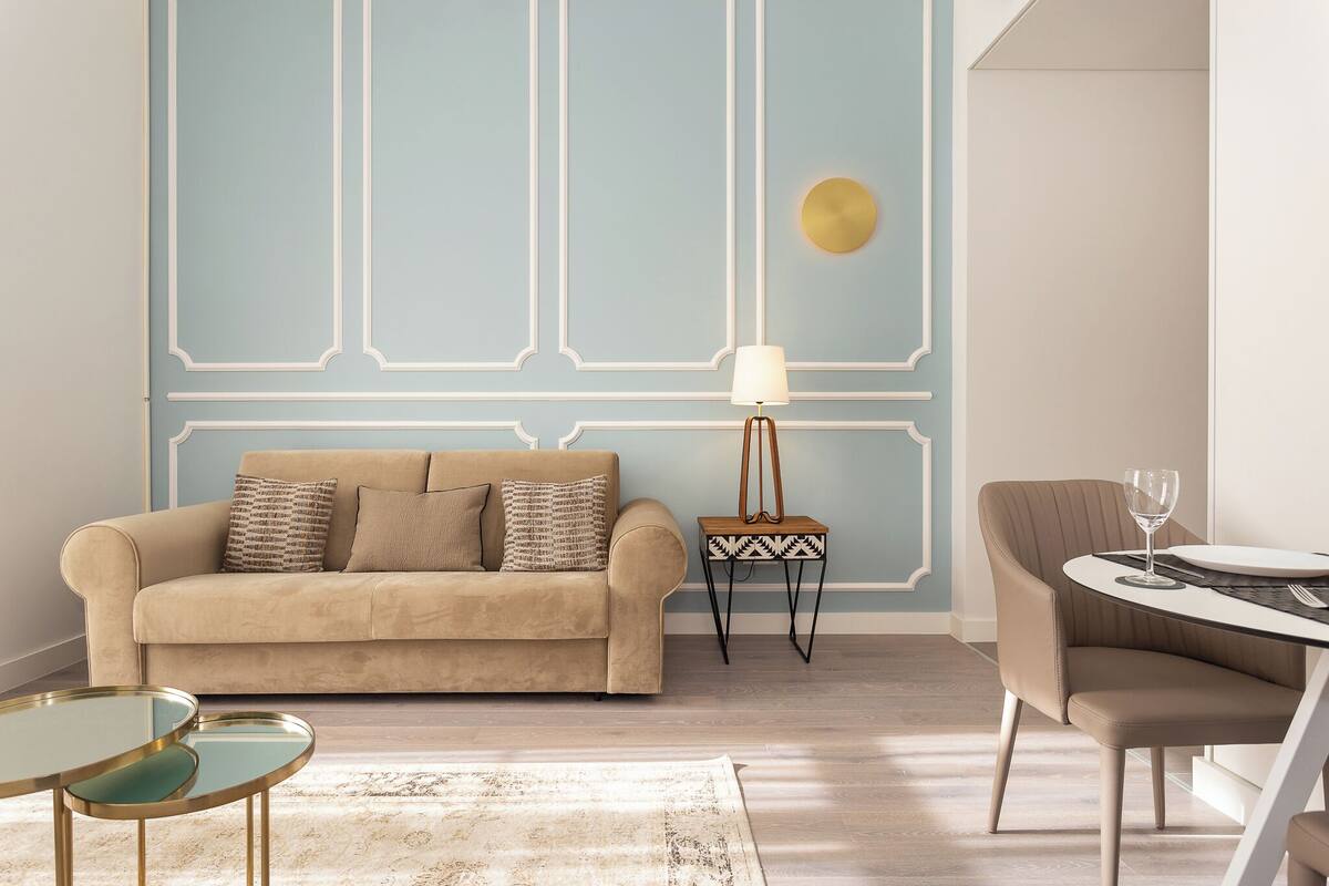 A living room with a beige sofa, a round coffee table, a side table with a lamp, and a dining area with a chair and a glass of wine.
