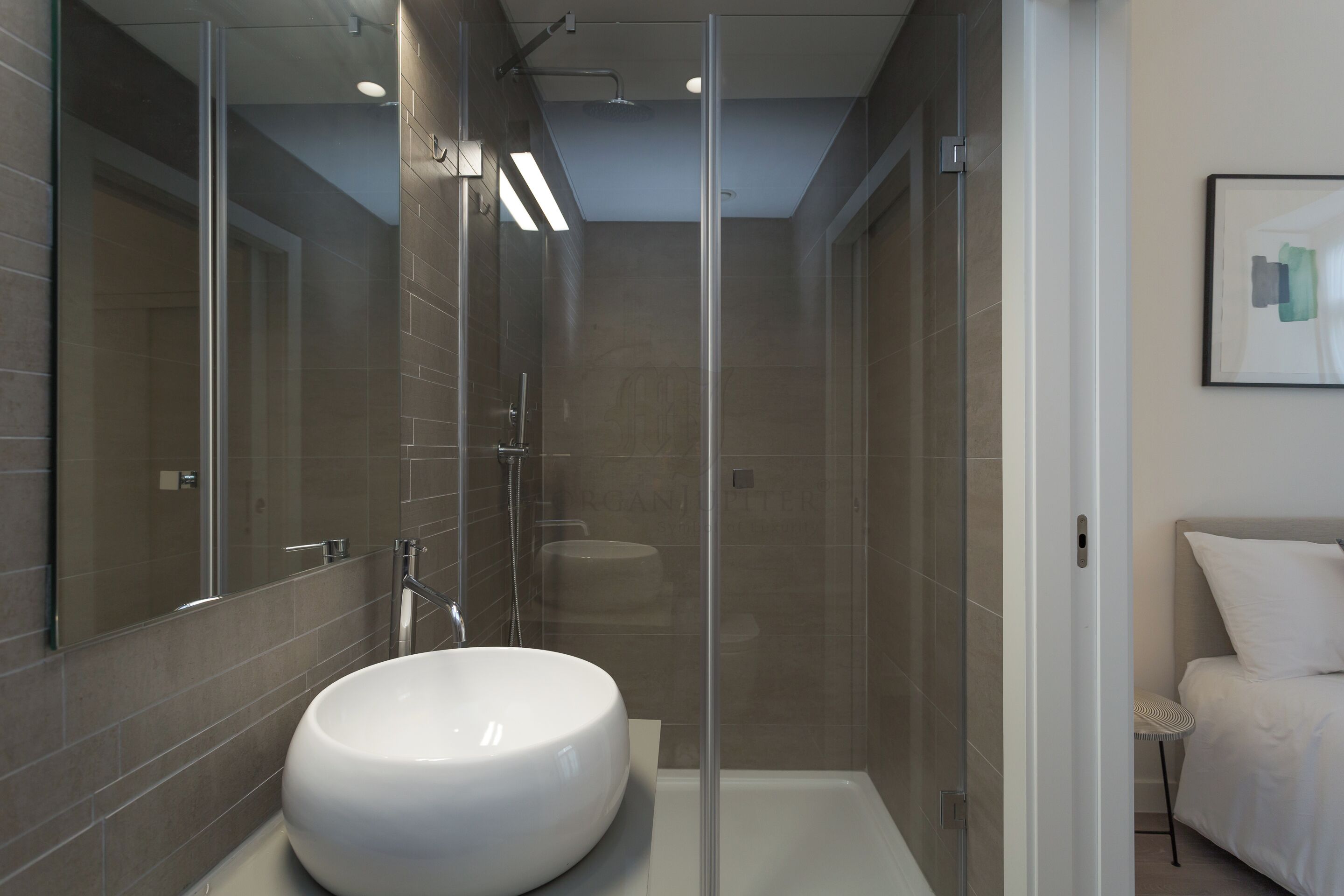 A modern bathroom with a large white sink, a glass-enclosed shower, and a bed with white bedding.