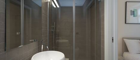 A modern bathroom with a large white sink, a glass-enclosed shower, and a bed with white bedding.