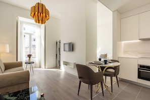 A modern living room with a sofa, a dining table with chairs, a TV mounted on the wall, and a kitchen area with cabinets and a stove.