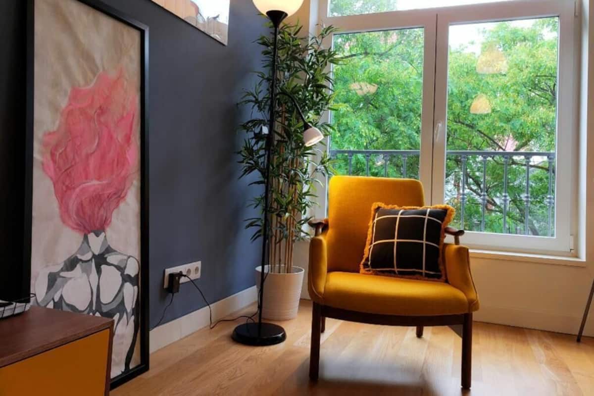 A room with a large window, a yellow armchair, a floor lamp, and a painting on the wall.