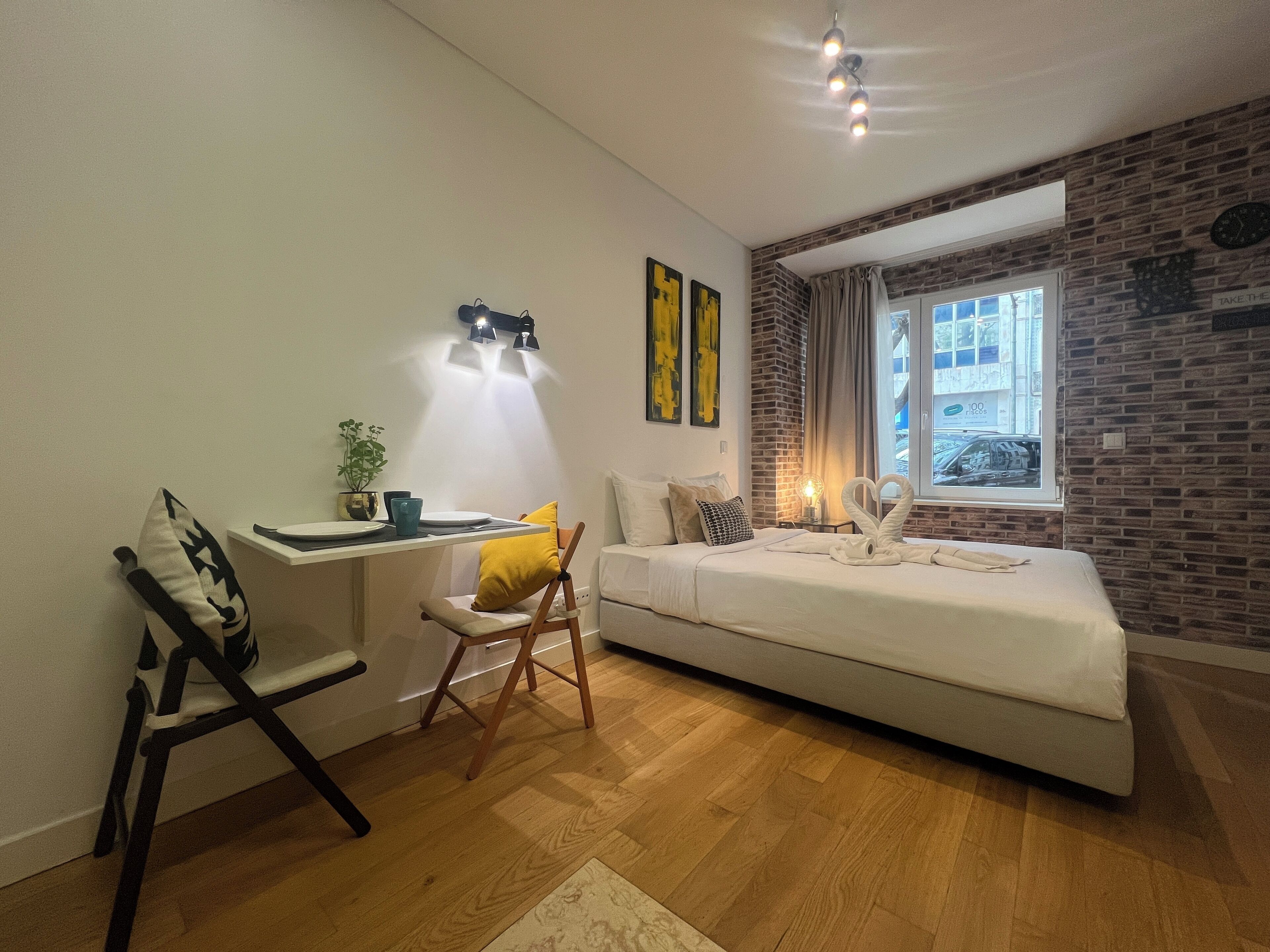 A modern bedroom with a brick accent wall, a large window, a bed, a desk with a chair, and a small table with a plant.