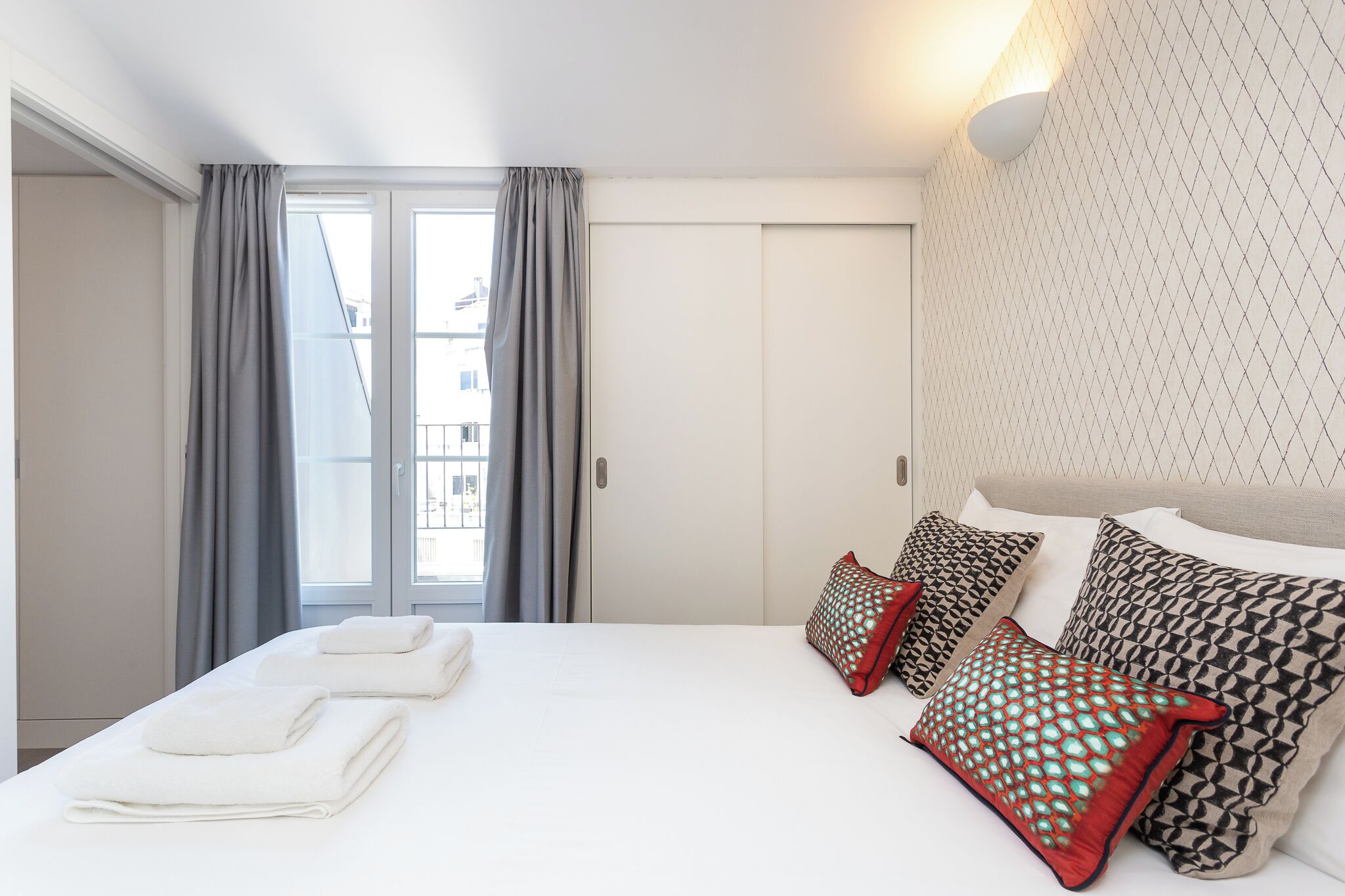 Una camera d'albergo con un letto, tende grigie, una testiera bianca e cuscini decorati.