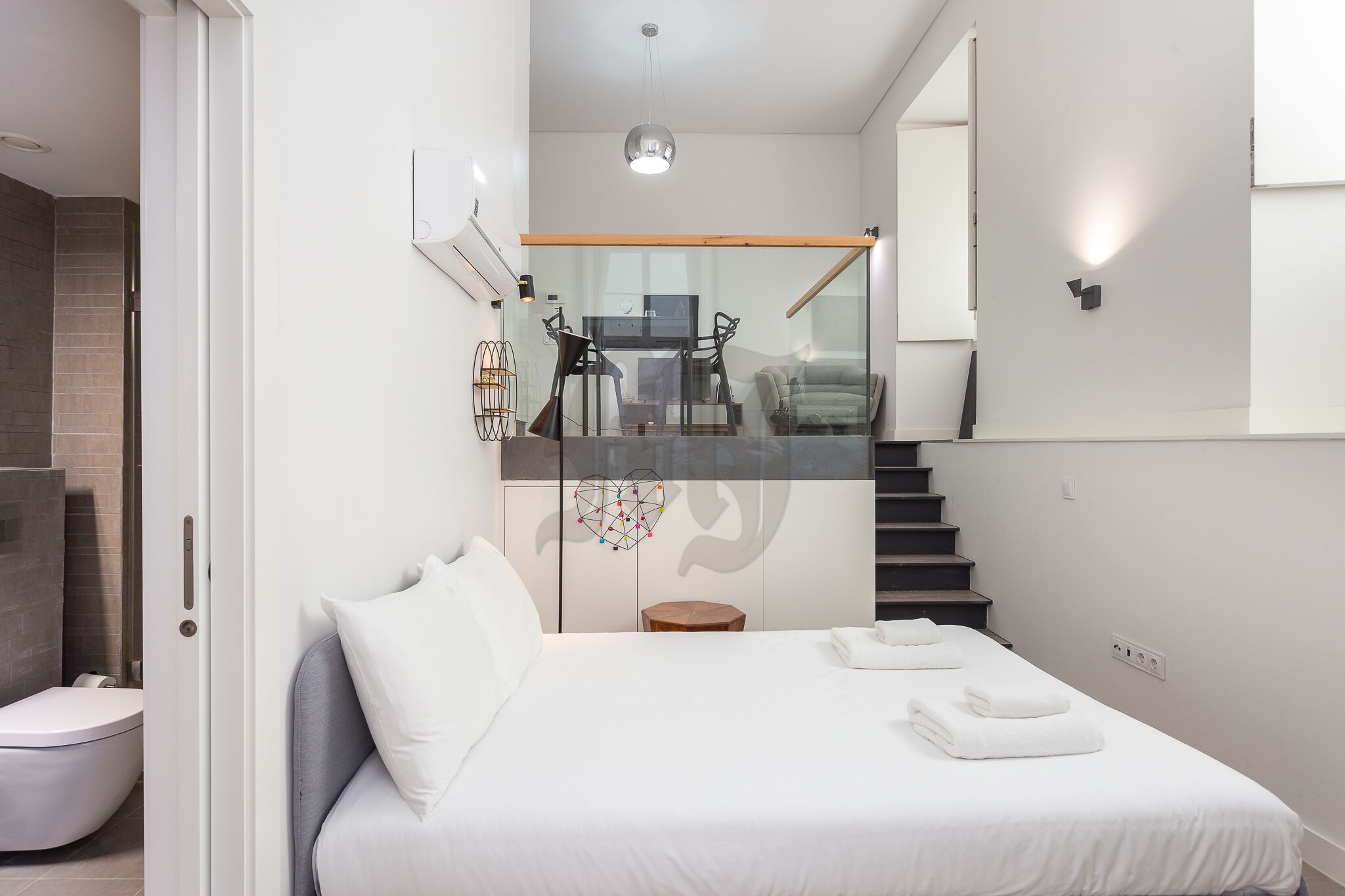 Quarto moderno com cama grande, divisória de vidro, escada e vista para uma área de refeições.