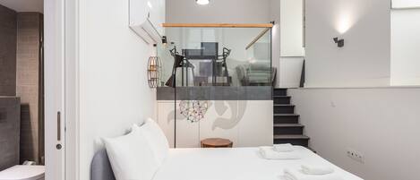 Quarto moderno com cama grande, divisória de vidro, escada e vista para uma área de refeições.