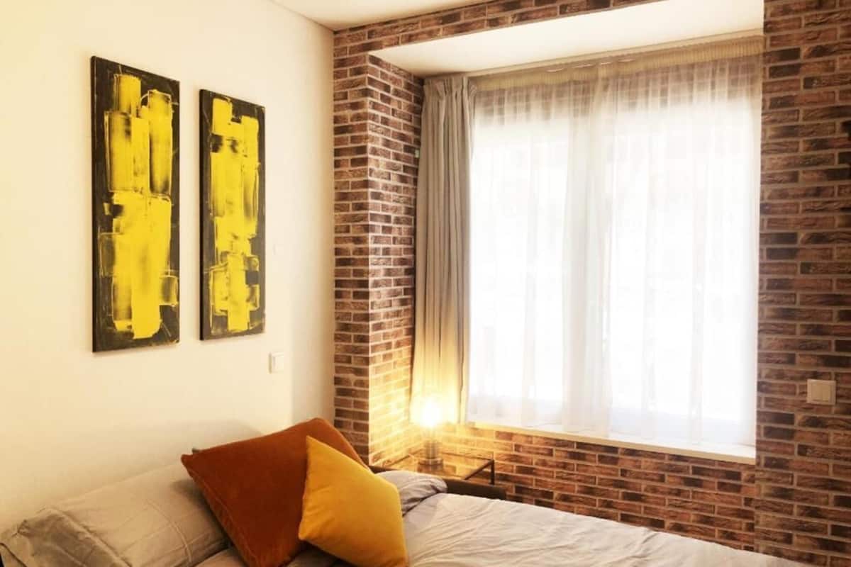 A bedroom with a brick wall, a bed with pillows, and a window with curtains.