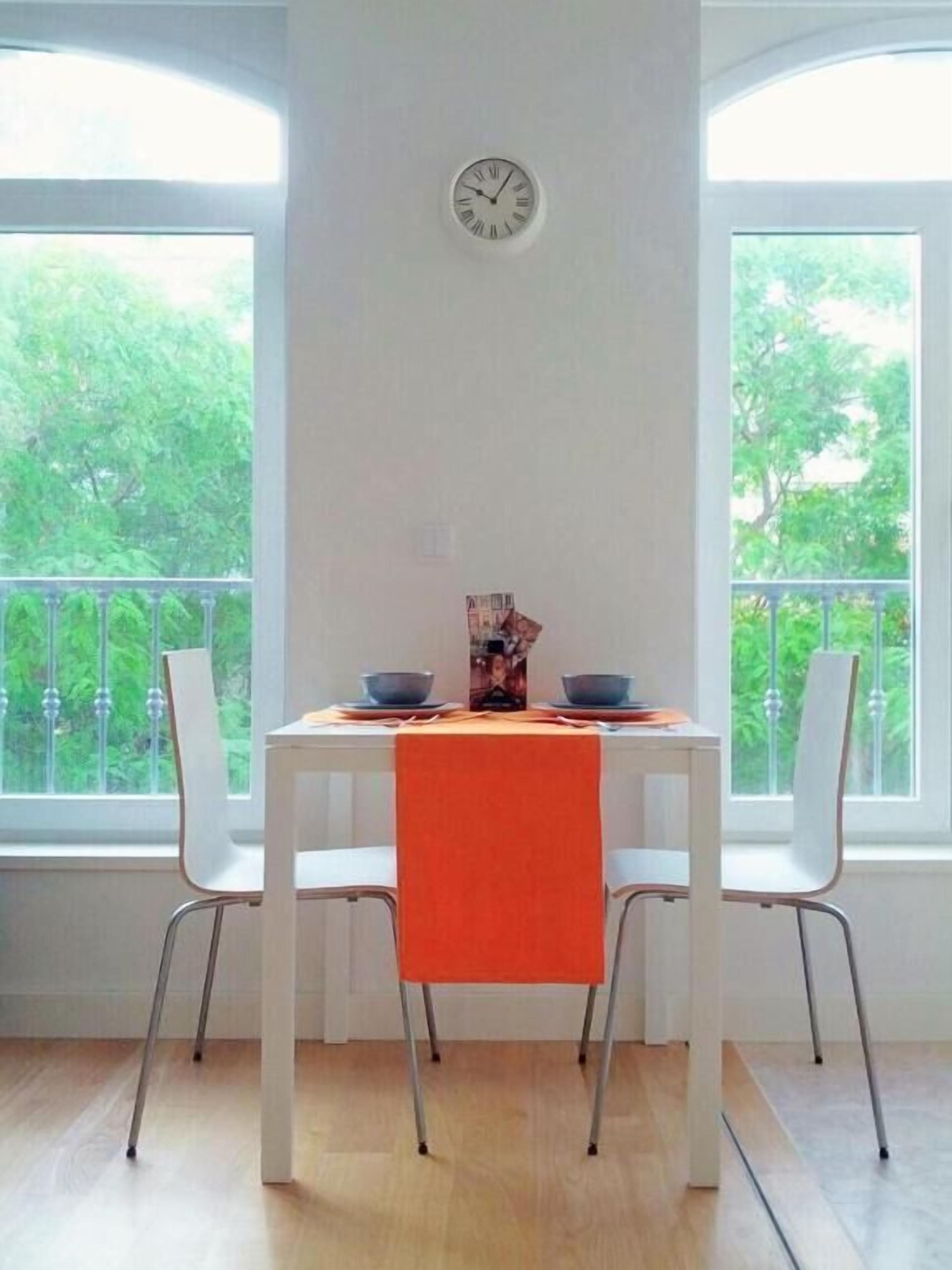 A dining area with a white table and chairs, a clock on the wall, and a view of greenery through large windows.