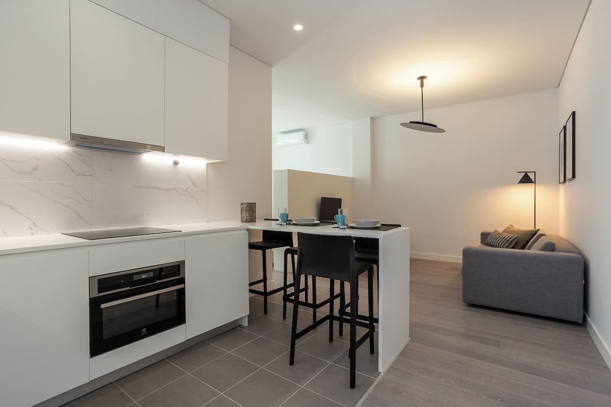 A modern kitchen with built-in appliances, a dining area with bar stools, and a grey sofa in a living space.