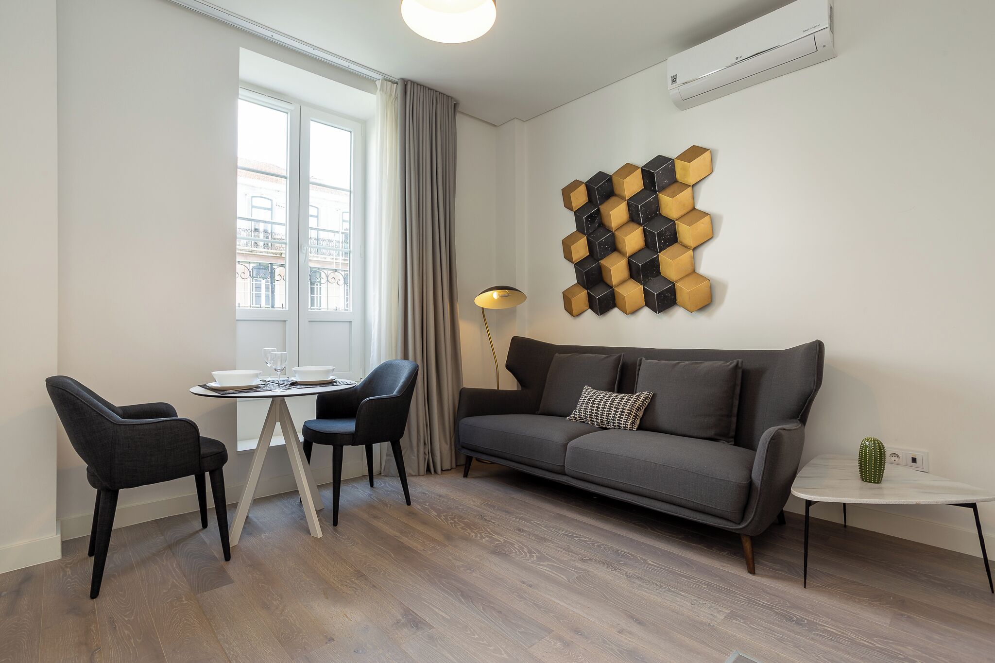 Una sala de estar moderna con un sofá gris, una mesa de comedor con dos sillas, un adorno en la pared y un pequeño cactus sobre una mesita auxiliar.