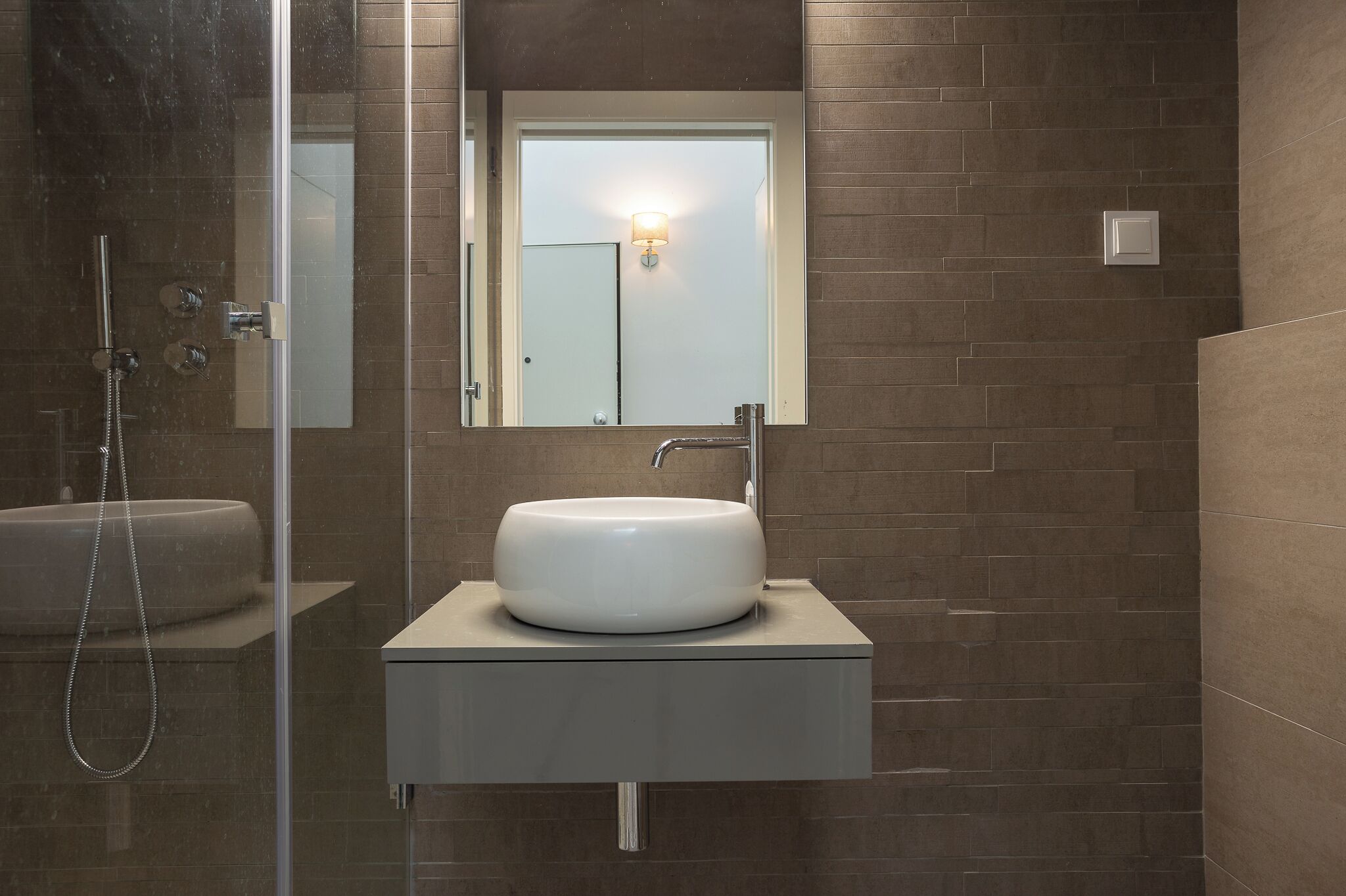 Un baño moderno con ducha envuelta en cristal, lavamanos blanco y espejo colgado en la pared.