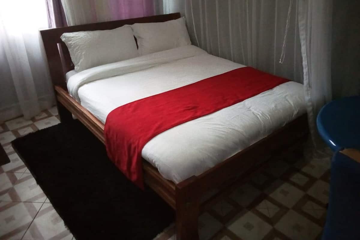Uma cama de solteiro com estrutura de madeira, roupa de cama branca e um cobertor vermelho. Há um tapete preto aos pés da cama e uma cadeira azul à direita.