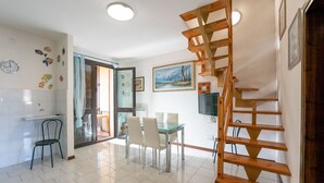 Een ruime woonkamer met een houten trap, een eettafel met stoelen, een keuken met spoelbak en een aan de muur bevestigde televisie.