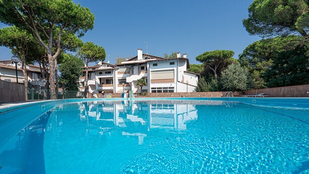 A swimming pool with clear blue water, surrounded by buildings and trees. - Spinnaker (Comacchio)