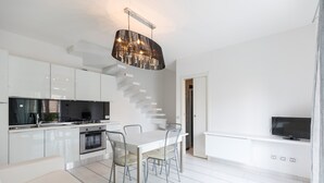 Una cocina moderna con armarios blancos, un comedor con mesa y sillas blancas, un televisor integrado y una escalera que sube a otro nivel.